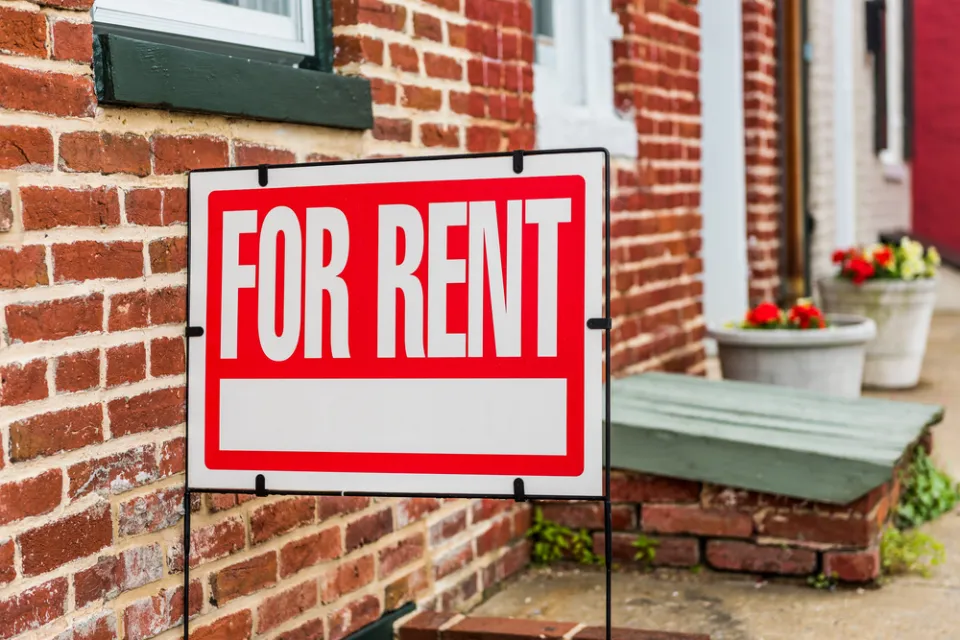 Red and white For Rent sign in front of brick building with flowerpots in background
